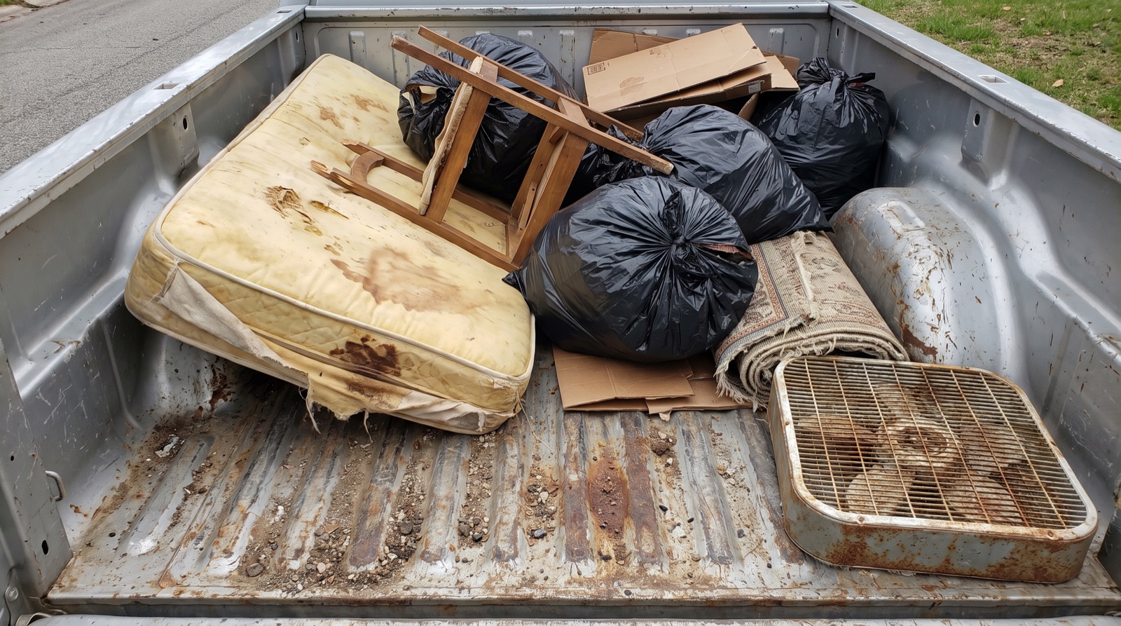 Illustrative AI-generated top-down image of a pickup truck bed loaded with a mattress, chair, trash bags and cardboard — not a TM Junk Removal job